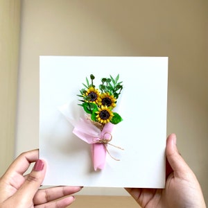 May include: A white card with a pink and brown paper bouquet of yellow sunflowers with green leaves.