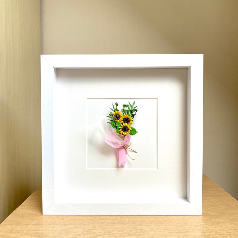 May include: A white frame with a white mat surrounding a small bouquet of yellow sunflowers with green leaves and a pink ribbon tied around the stems.