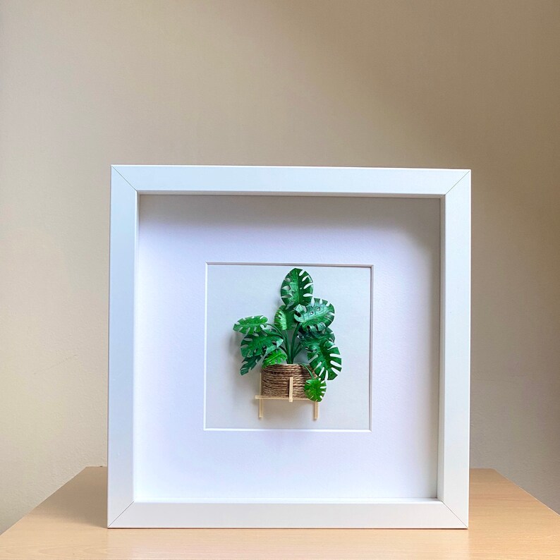 May include: A white framed picture with a green paper plant in a brown pot on a small wooden stand. The plant is a monstera deliciosa.