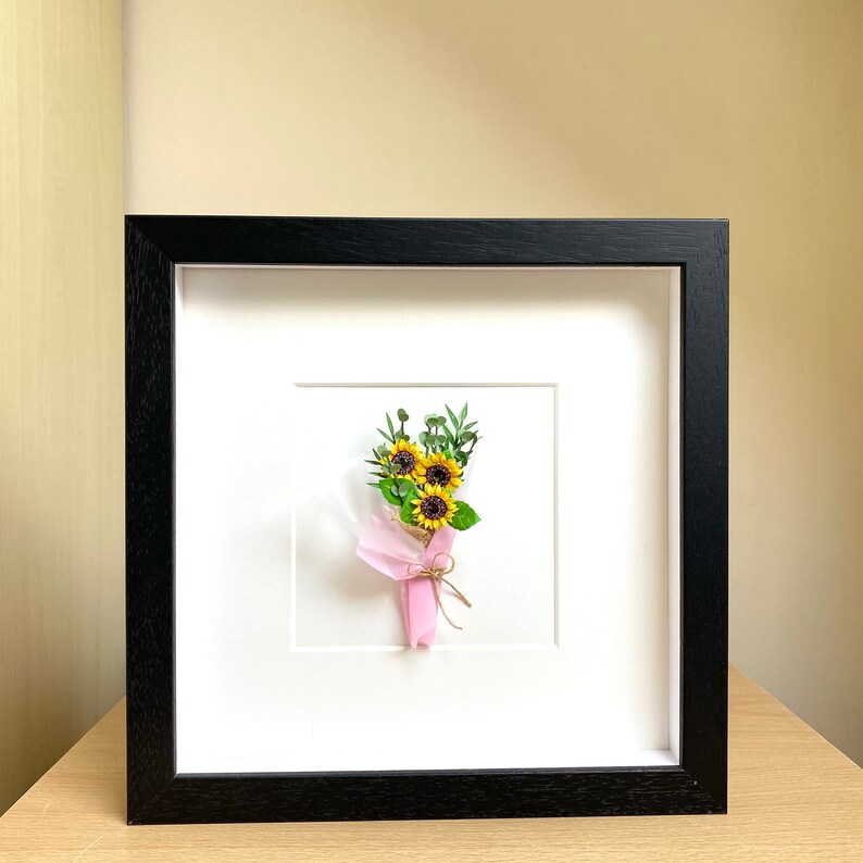 May include: A framed artwork featuring a small bouquet of paper sunflowers tied with a pink ribbon and twine. The sunflowers are yellow with brown centers and green leaves. The artwork is framed in a black frame.