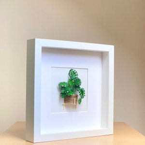May include: White framed picture with a green paper plant in a woven basket on a small wooden stand. The plant is a monstera deliciosa.