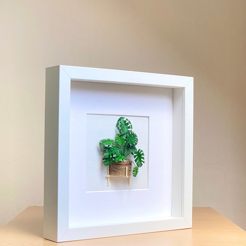 May include: White framed picture with a green paper plant in a woven basket on a small wooden stand. The plant is a monstera deliciosa.