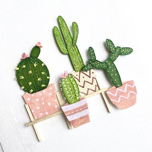 May include: Three paper cacti in pink pots with white polka dots, sitting on a wooden shelf. The cacti are green and have a variety of shapes and sizes.