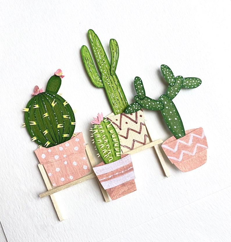 May include: Three paper cacti in pink pots with white polka dots, sitting on a wooden shelf. The cacti are green and have a variety of shapes and sizes.