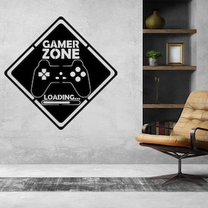 Puede incluir: Adhesivo de pared Gamer Zone en blanco y negro con un diseño de mando de videojuegos y la palabra "LOADING...". El adhesivo tiene forma de diamante y está montado en una pared gris. Una silla de cuero marrón y una lámpara de pie están en el fondo.