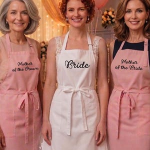 May include: Three aprons worn by individuals: two light pink aprons with the text Mother of the Groom and Mother of the Bride, and one white apron in the center with the text Bride. Each apron features a tie waist and a front pocket.