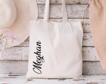 Puede incluir: Un bolso de tela color crema con dos asas para el hombro y el nombre Meghan impreso verticalmente en una fuente cursiva negra en la parte frontal. El bolso tiene una forma rectangular y está hecho de un material de tela sencillo.