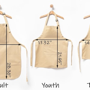 Puede incluir: Tres delantales beige etiquetados como Adult, Youth y Toddler, colgados en perchas de madera. El delantal de adulto mide 54 cm de ancho por 71,88 cm de largo. El delantal juvenil mide 43,99 cm de ancho por 54,99 cm de largo y el de ni&ntilde;o 38 cm por 44,98 cm.