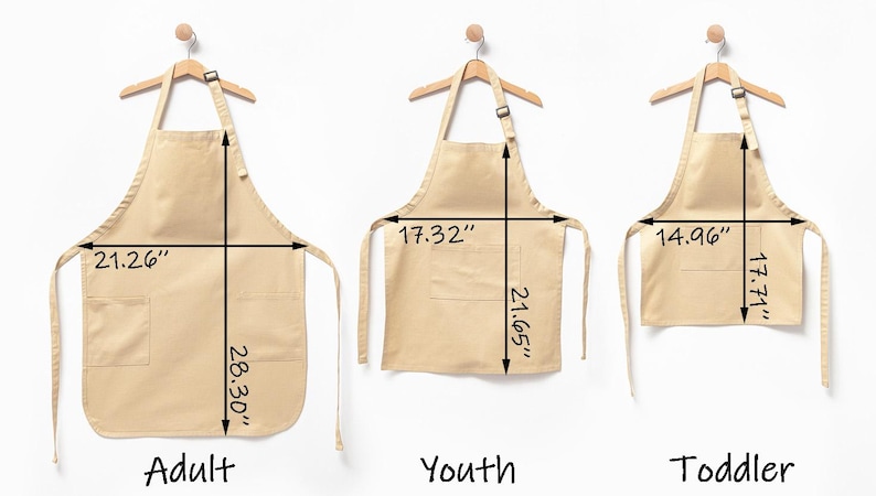 Puede incluir: Tres delantales beige etiquetados como Adult, Youth y Toddler, colgados en perchas de madera. El delantal de adulto mide 54 cm de ancho por 71,88 cm de largo. El delantal juvenil mide 43,99 cm de ancho por 54,99 cm de largo y el de ni&ntilde;o 38 cm por 44,98 cm.