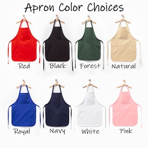 Puede incluir: Ocho delantales colgados en perchas de madera, con el texto Apron Color Choices. Los colores son rojo, negro, verde bosque, natural, azul real, azul marino, blanco y rosa. Cada delantal tiene una cinta para el cuello y dos bolsillos.