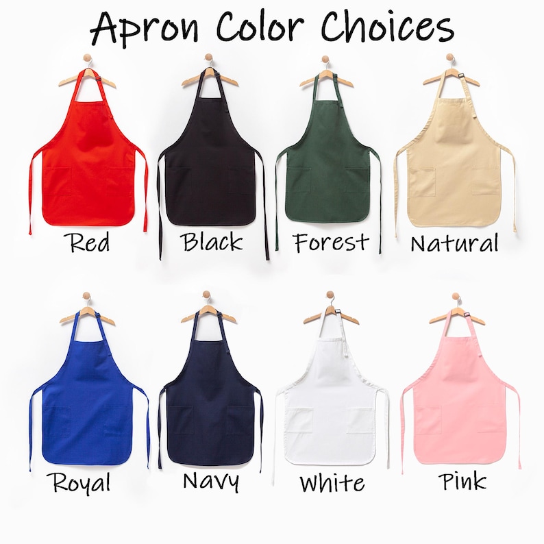 Puede incluir: Ocho delantales colgados en perchas de madera, con el texto Apron Color Choices. Los colores son rojo, negro, verde bosque, natural, azul real, azul marino, blanco y rosa. Cada delantal tiene una cinta para el cuello y dos bolsillos.