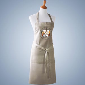 Puede incluir: Un delantal beige con un gr&aacute;fico circular en el pecho con el texto YOUR LOGO HERE en negro y naranja. El delantal tiene una correa para el cuello, un lazo en la cintura y un bolsillo frontal grande, exhibido en un maniqu&iacute; blanco sobre fondo azul.
