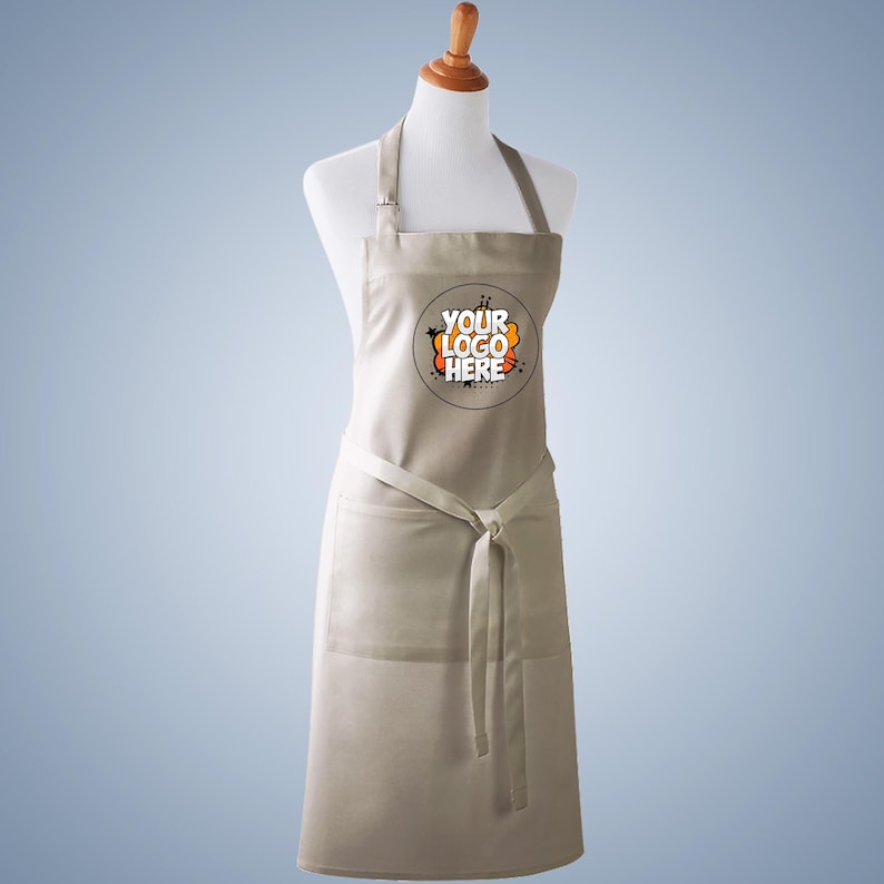 Puede incluir: Un delantal beige con un gr&aacute;fico circular en el pecho con el texto YOUR LOGO HERE en negro y naranja. El delantal tiene una correa para el cuello, un lazo en la cintura y un bolsillo frontal grande, exhibido en un maniqu&iacute; blanco sobre fondo azul.