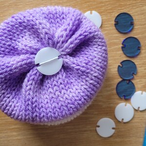 Könnte beinhalten: Eine lila und weiße gehäkelte Mütze mit einem weißen Knopf oben. Die Mütze ist umgeben von blauen und weißen Plastik-Maschenmarkern und einer blauen Häkelnadel.
