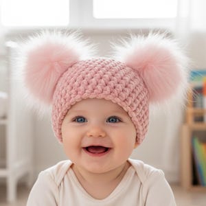 May include: A baby wearing a pink knitted hat with two large, fluffy pom-poms. The hat is a soft, pastel pink color. The baby is smiling, with blue eyes and a white onesie.