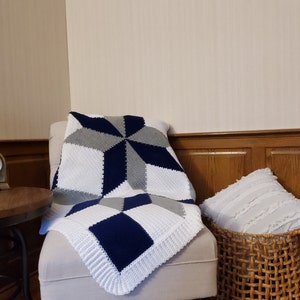 May include: A white, gray, and navy blue crocheted throw blanket with a starburst pattern. The blanket is draped over a white armchair with a white pillow in a wicker basket.