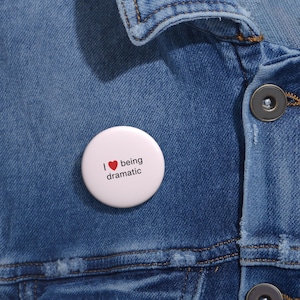 Può includere: Un bottone bianco con la scritta "I ❤️ being dramatic" in nero, con un simbolo a forma di cuore rosso. Il bottone è appuntato su una giacca di jeans blu.