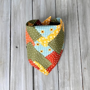 Peut inclure: Un bandana triangulaire pour animal avec un motif patchwork composé de divers tissus. Les motifs incluent du vert olive à pois blancs, du bleu clair à carreaux blancs, du jaune avec de petites tournesols et un tissu orange vif à pois blancs.