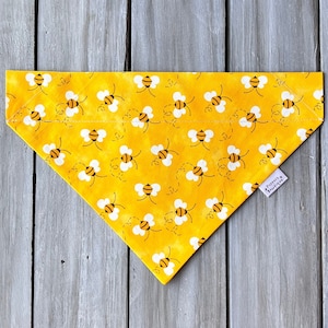 May include: Yellow dog bandana featuring a pattern of flying bees. The bandana has a white tag with the text "Pawsitive Boutique" on it.