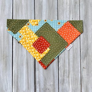 Op de afbeelding: Een driehoekige bandana voor huisdieren met een patchworkontwerp van verschillende stoffen vierkanten. De patronen omvatten olijfgroen met witte stippen, feloranje met witte stippen, lichtblauw met een ruitpatroon en geel met witte bloemen.