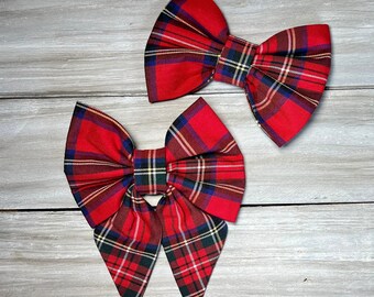 May include: Two red plaid fabric bows with a classic tartan pattern. The bows feature a mix of red, green, blue, and yellow lines. One bow has long, pointed tails. The bows are set against a light gray wooden background.