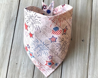 May include: A light pink dog bandana with a patriotic fireworks and star pattern. The bandana features red, white, and blue stars and fireworks. It is secured with a blue button.