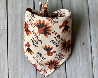 May include: A beige bandana with a repeating pattern of turkeys and the text "Gobble 'til you Wobble!" in a brown, orange, and black color scheme.
