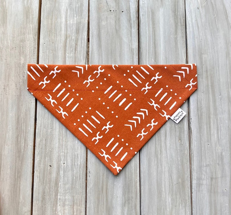 May include: A rust-colored dog bandana with a white geometric pattern. The bandana has a white tag that says "Yours & Mine".