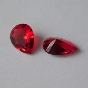 Könnte beinhalten: Zwei rote birnenförmige facettierte Glasperlen auf weißem Hintergrund.