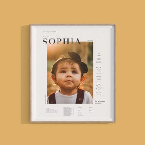 May include: Framed print featuring a portrait of a young child named Sophia. The print includes the child's name in large text, along with other details such as birth date, weight, and length. The frame is a light wood color, and the background is a warm, neutral tone.