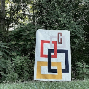 May include: A white quilt with a geometric design featuring overlapping squares in red, yellow, black, and brown. The quilt is displayed outdoors against a backdrop of green trees.