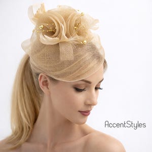 May include: A beige fascinator with a floral design and gold bead accents. The headpiece features a mesh base and a large, layered flower. The text "AccentStyles" is visible in the image.
