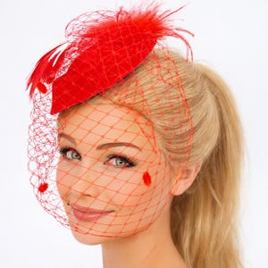 May include: A vibrant red fascinator with a matching veil. The headpiece features a small, rounded base adorned with feathers and a delicate net veil that drapes over the face. The accessory is designed for special occasions.