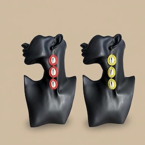 Peut inclure: Une paire de longues boucles d'oreilles pendantes. Une boucle d'oreille est rouge avec des accents blancs, l'autre est jaune avec des accents blancs. Chaque boucle d'oreille comporte trois éléments de forme ovale. Les boucles d'oreilles sont exposées sur des têtes de mannequin noires.