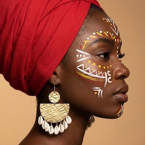 Peut inclure: Profil d'une personne portant un foulard rouge et des boucles d'oreilles dorées. Les boucles d'oreilles ont un motif en demi-cercle en or martelé avec des accents de coquillages blancs. Le visage est orné de peinture faciale blanche, jaune et rouge.