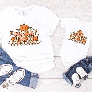 May include: White t-shirts with the text "Mama Pumpkin" and "Mini Pumpkin" in orange and brown lettering with a pumpkin graphic. The shirts are paired with jeans and white sneakers.