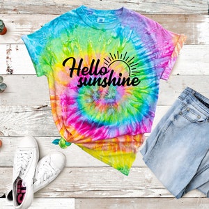 May include: A tie-dye t-shirt with a rainbow swirl pattern and the text "Hello sunshine" in black. The shirt is laid out on a wooden surface with a pair of white sneakers and blue jeans.