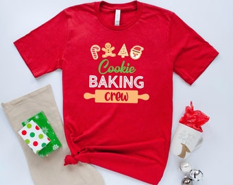 Pode incluir: Camiseta vermelha com um gráfico branco que diz "Cookie Baking Crew" e apresenta um homem de gengibre, um bastão de caramelo, uma árvore de Natal e Papai Noel.