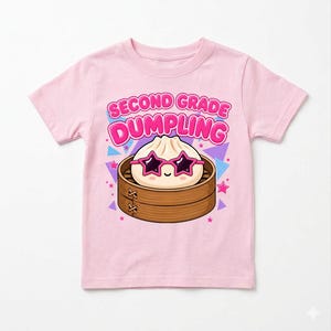 May include: A light pink t-shirt featuring a graphic of a cartoon dumpling wearing star-shaped sunglasses inside a bamboo steamer basket. The text above the graphic reads SECOND GRADE DUMPLING in bold pink letters, surrounded by small stars and triangles.