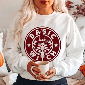 Könnte beinhalten: Weißer Pullover mit einem roten Grafikdruck einer Hexe mit Hut und dem Text "BASIC WITCH" in einem Kreis.