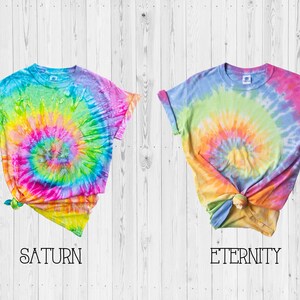 May include: Two tie-dye t-shirts with a rainbow swirl pattern. The shirt on the left is labeled "SATURN" and the shirt on the right is labeled "ETERNITY".