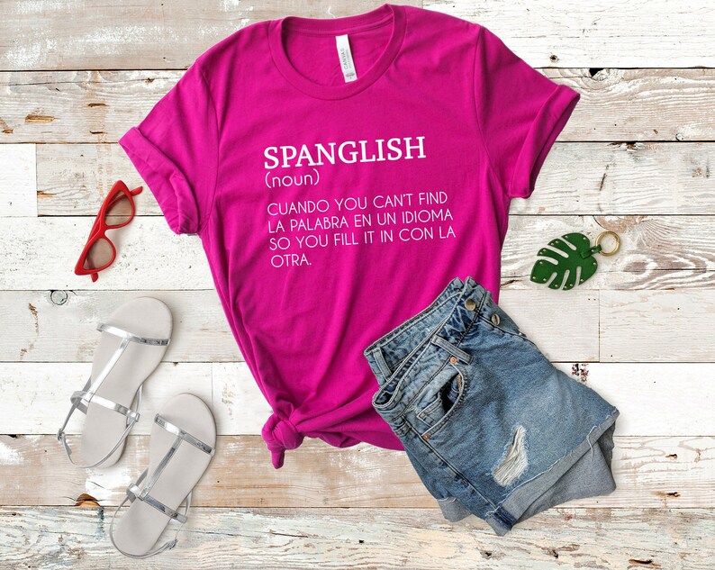 Pode incluir: Uma camiseta rosa com a palavra "Spanglish" definida como "Quando voc&ecirc; n&atilde;o encontra a palavra em um idioma, voc&ecirc; a completa com o outro."