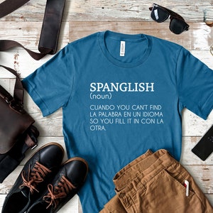 Pode incluir: Uma camiseta azul com texto branco que diz "SPANGLISH (nome) CUANDO YOU CAN'T FIND LA PALABRA EN UN IDIOMA SO YOU FILL IT IN CON LA OTRA."
