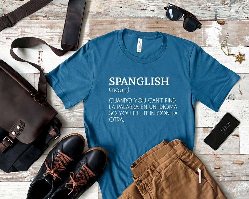 Pode incluir: Uma camiseta azul com texto branco que diz "SPANGLISH (nome) CUANDO YOU CAN'T FIND LA PALABRA EN UN IDIOMA SO YOU FILL IT IN CON LA OTRA."