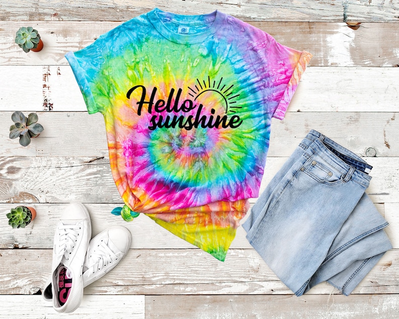 May include: A tie-dye t-shirt with a rainbow swirl pattern and the text "Hello sunshine" in black. The shirt is laid out on a wooden surface with a pair of white sneakers and blue jeans.
