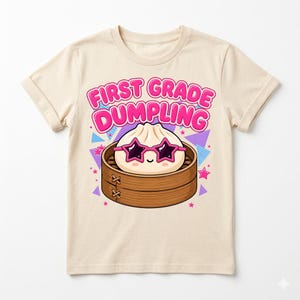 May include: A cream-colored t-shirt featuring a graphic of a cartoon dumpling wearing pink star-shaped sunglasses inside a bamboo steamer basket. The text above the dumpling reads FIRST GRADE DUMPLING in bold pink letters, surrounded by colorful geometric shapes.
