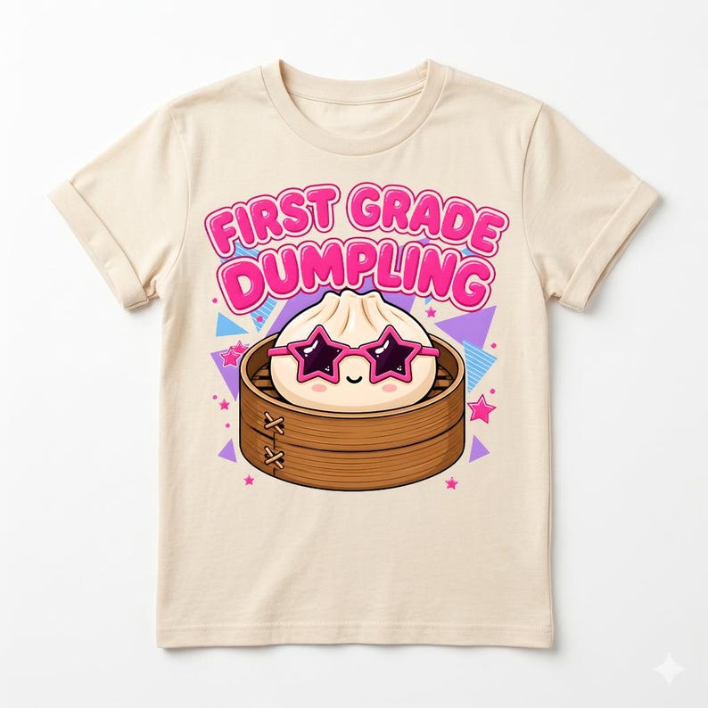 May include: A cream-colored t-shirt featuring a graphic of a cartoon dumpling wearing pink star-shaped sunglasses inside a bamboo steamer basket. The text above the dumpling reads FIRST GRADE DUMPLING in bold pink letters, surrounded by colorful geometric shapes.