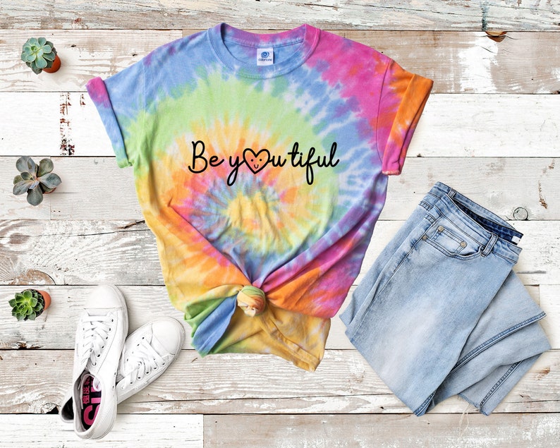May include: A tie-dye t-shirt with a rainbow swirl pattern. The shirt has the text "Be you tiful" with a heart in place of the "o". The shirt is paired with light blue jeans and white sneakers.