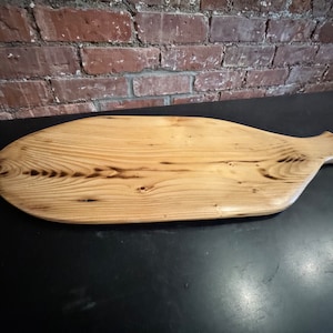 May include: A wooden serving board with a natural finish. The board is oval-shaped with a handle on one end. The wood grain is visible, and there are dark burn marks. The board is approximately 20 inches long.