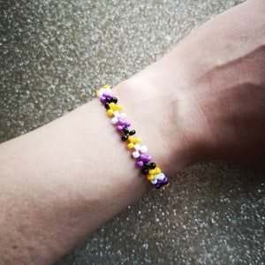 May include: A beaded bracelet with alternating purple, yellow, white and black beads. The bracelet is worn on a person's wrist.